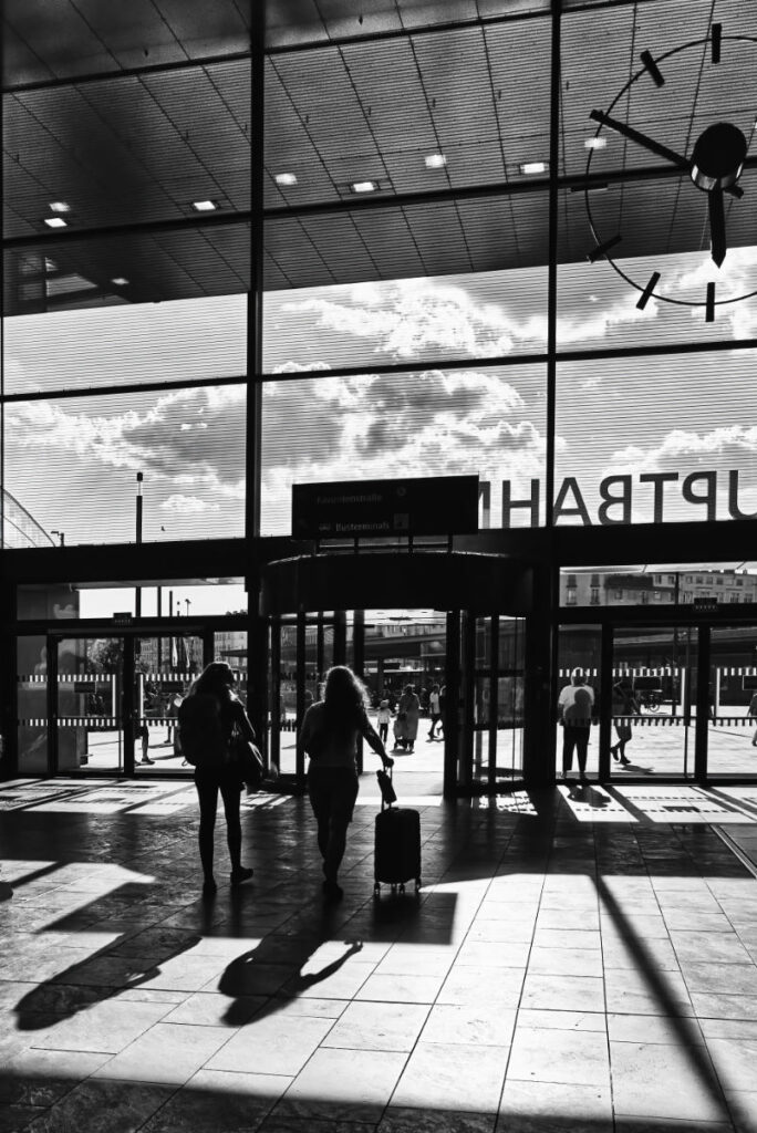 Zwei Personen mit Koffer als Silhouetten in der Bahnhofshalle, die große Bahnhofsuhr ist im Bild nur angeschnitten