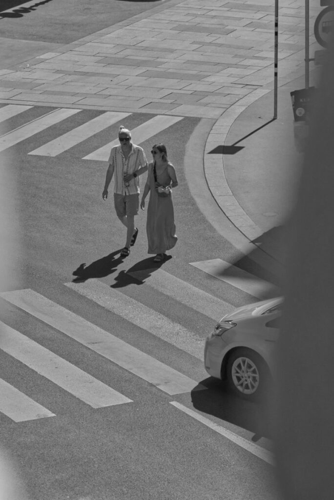 Ein Paar überquert von oben fotografiert einen Zebrastreifen, lange Schatten auf dem Asphalt