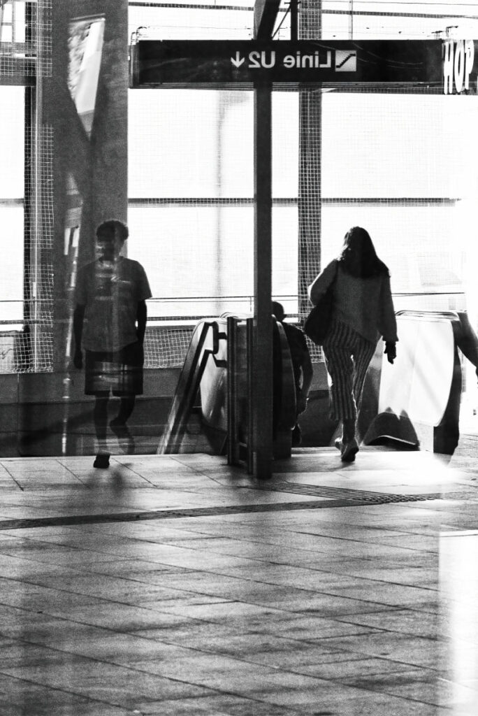Doppelbelichtungsartige Spiegelung einer Person neben einer klar erkennbaren Silhouette an einer Rolltreppe