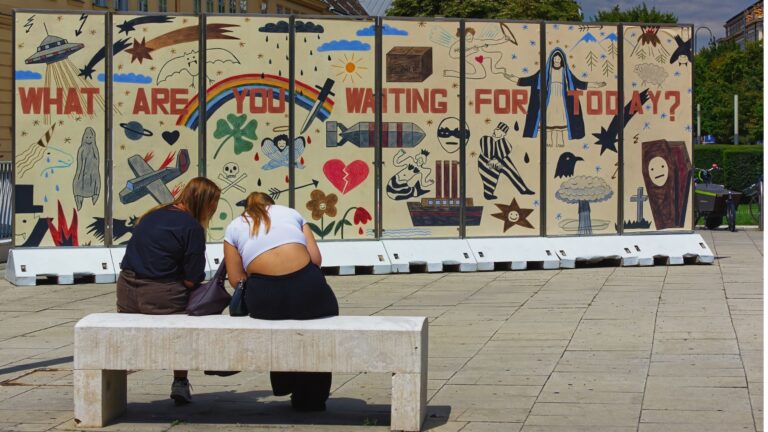 Zwei Frauen sitzen von hinten fotografiert auf einer Bank vor einer bunt bemalten Wand mit der Aufschrift "What are you waiting for today"