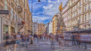 Menschen gehen an einem sonnigen Tag unscharf über den Graben in Wien vorbei, im Hintergrund die Pestsäule
