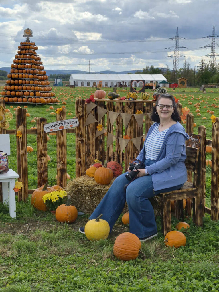 Barbara mit Kamera auf einem Holzsessel am Pumpkin Patch, dahinter Kürbisfeld, Kürbispyramide und weißes Zelt