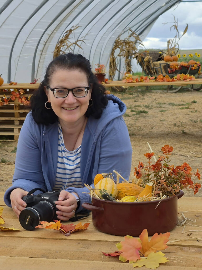 Barbara mit Kamera an einem Holztisch mit Zierkürbissen und Herbstlaub in einer Scheune