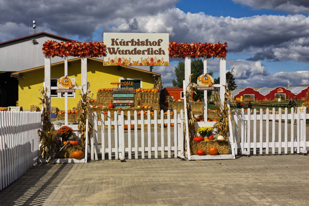 Eingangstor zum Kürbishof Wunderlich mit Holzschild und Herbstlaub, dahinter Strohballen mit Kürbissen und rote Scheunen